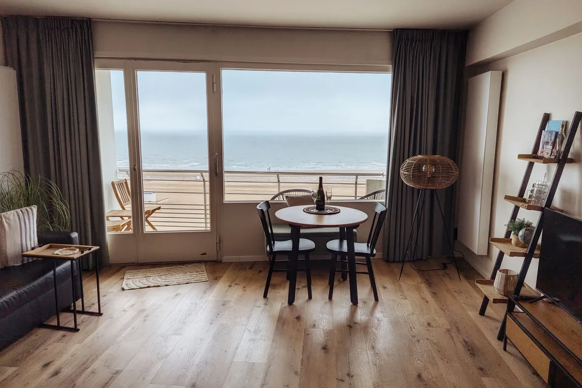 Holiday flat living room with a round table for two, balcony doors onto the sea and the television switched off on the right
