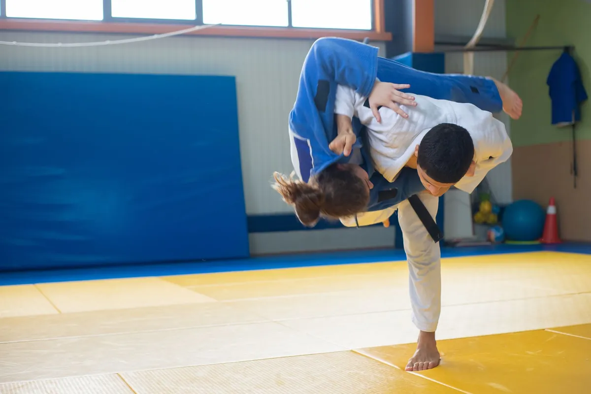 Tatami de un dojo con dos practicantes de judo en plena proyección, una de blanco con cinturón negro y otra de azul
