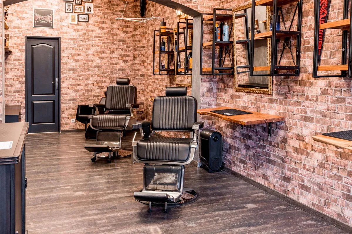 Barbershop interior with two black leather chairs on a wood floor, exposed brick walls and product shelves between gilt mirrors