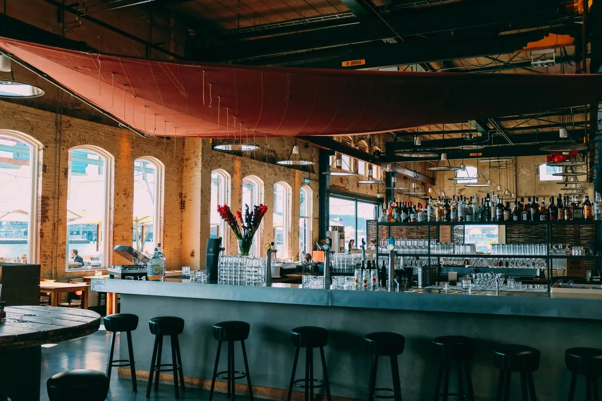 Balcão comprido de bar com uma fileira de banquetas pretas, a prateleira de garrafas ao fundo e um toldo vermelho esticado sob o teto industrial