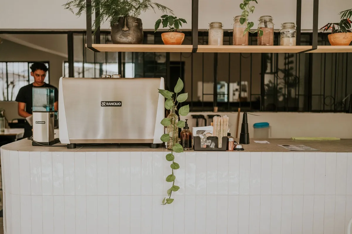 Balcão de cafeteria revestido de azulejo branco com a máquina de espresso e o moedor, potes e plantas na prateleira de cima e um barista ao fundo