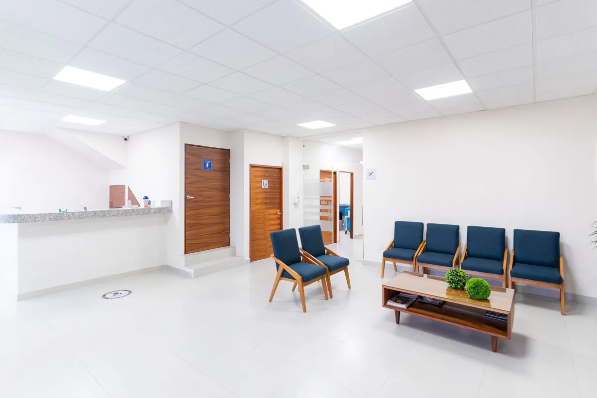 Sala de espera de una clínica con el mostrador de recepción a la izquierda, cuatro butacas azules alineadas contra la pared y una mesa baja con plantas