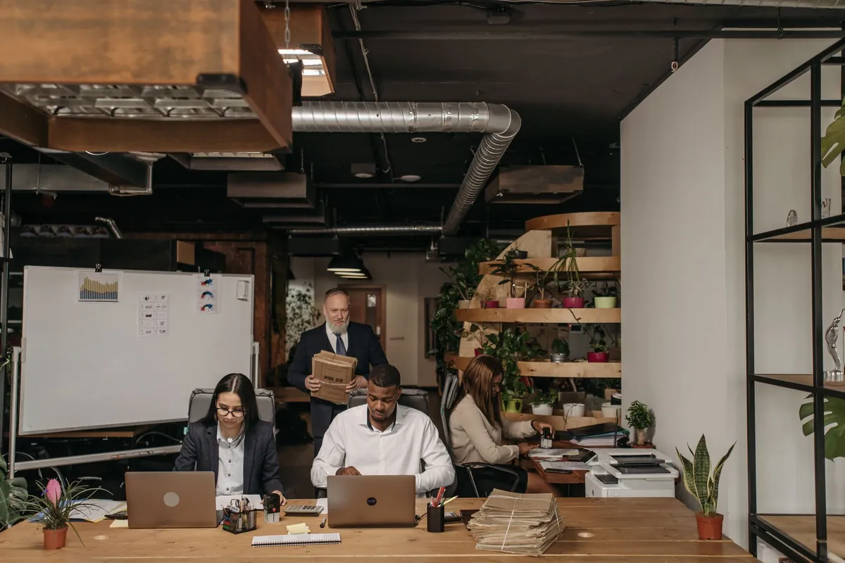 Spazio di coworking con un lungo tavolo di legno, portatili aperti, una lavagna bianca con grafici stampati e scaffali di piante sul fondo