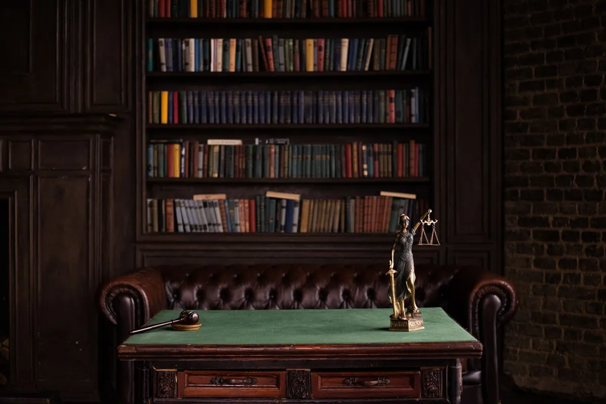 Law office with the law library behind, a leather chesterfield sofa and a green-baize desk holding a gavel and a figure of Justice
