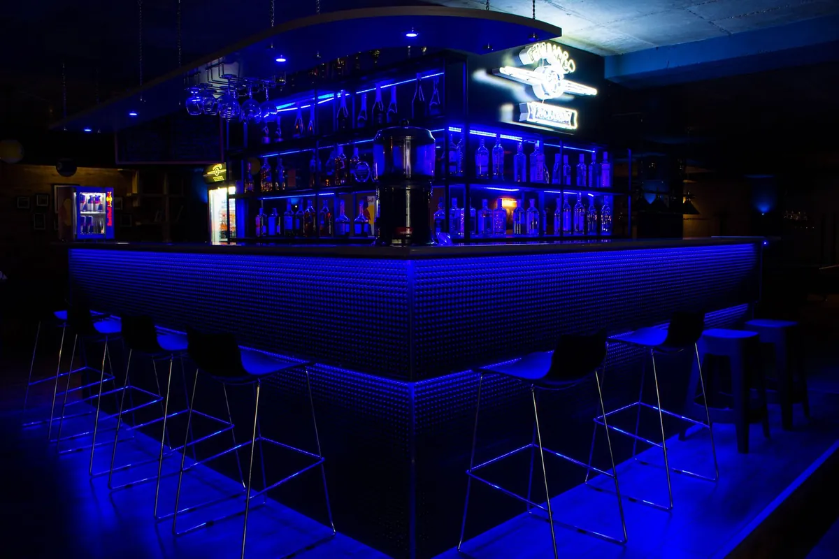 Bancone di una discoteca in penombra con il frontale illuminato di blu, gli sgabelli alti in fila e la scaffalatura delle bottiglie retroilluminata