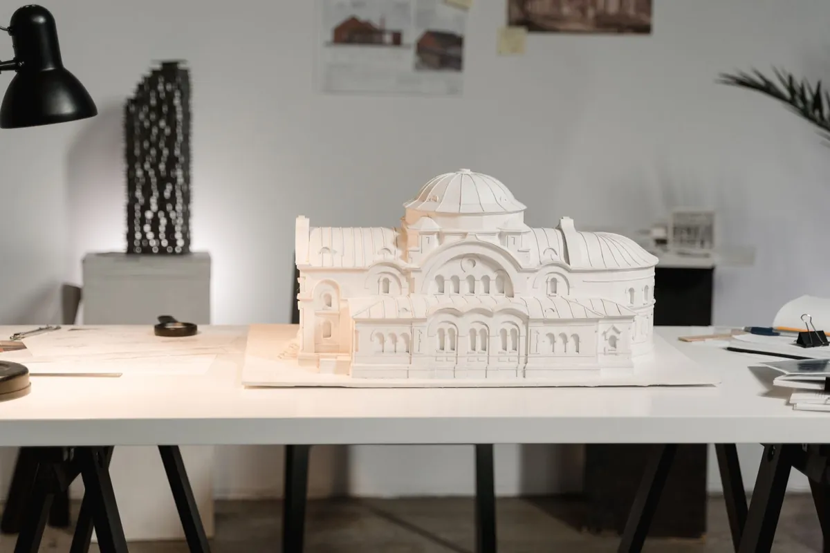 White scale model of a domed building on the trestle table of an architecture studio, with the desk lamp and drawings pinned behind