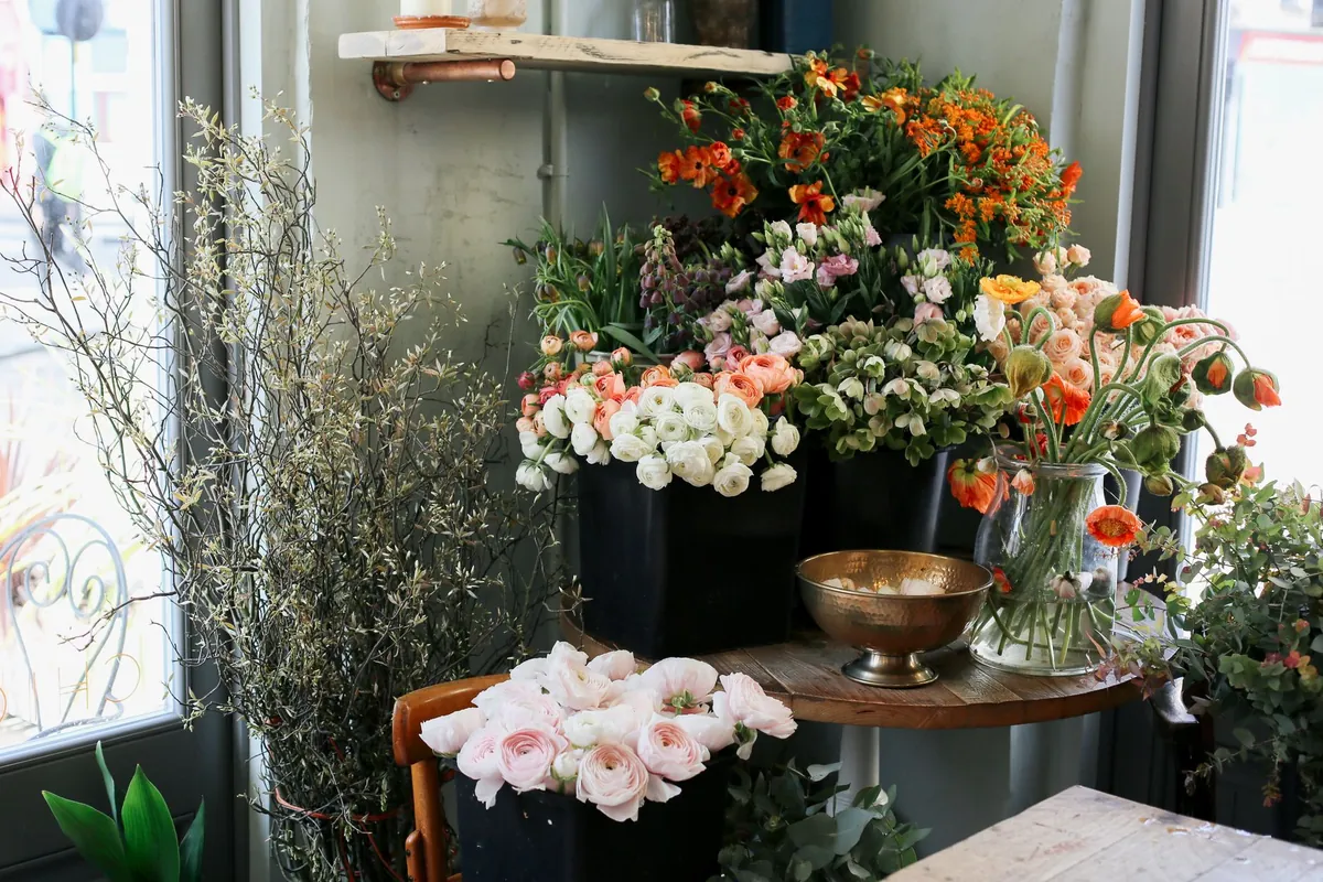 Ecke eines Blumenladens am Schaufenster mit Eimern voller Ranunkeln und Mohn, einer Messingschale und trockenen Zweigen an der Scheibe