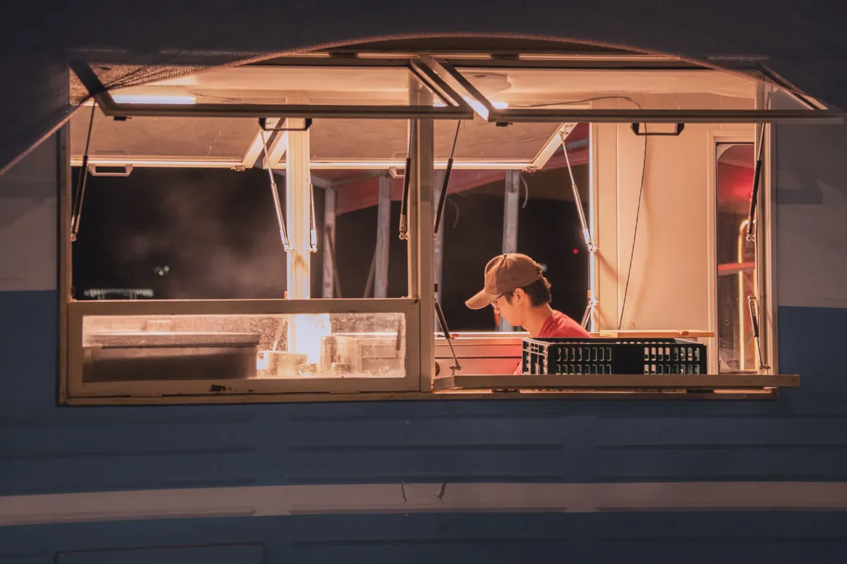 Fenêtre de service d'un food truck la nuit, éclairée de l'intérieur, avec quelqu'un en casquette et une caisse sur le comptoir