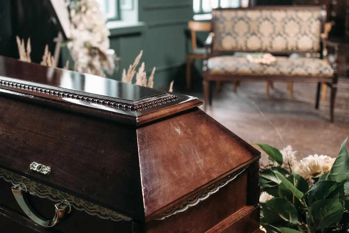 Sala de velório com o caixão de madeira fechado em primeiro plano, flores brancas dos lados e um banco estofado ao fundo