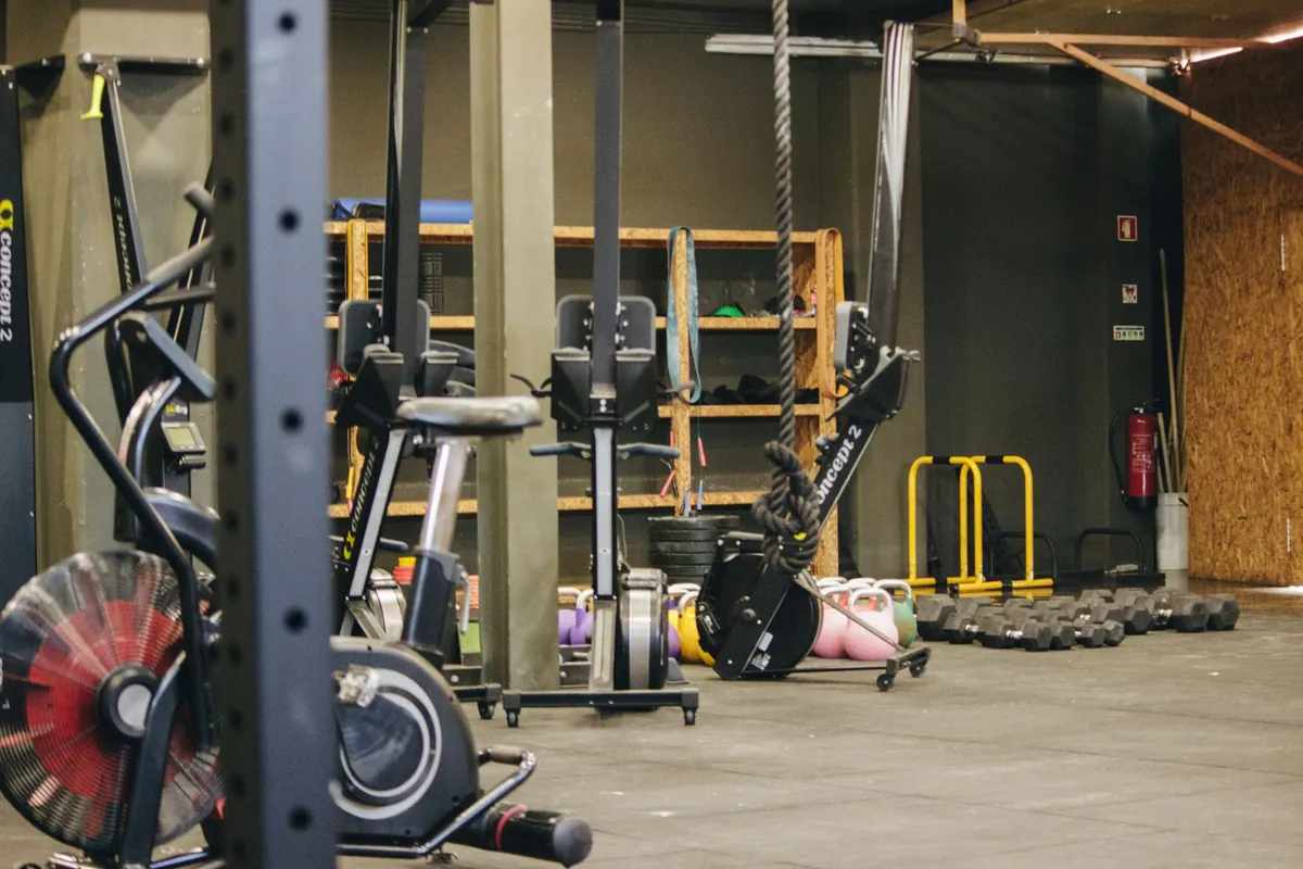 Área de treino com remos ergométricos, air bike, kettlebells enfileirados e um rack de halteres junto à parede de madeira