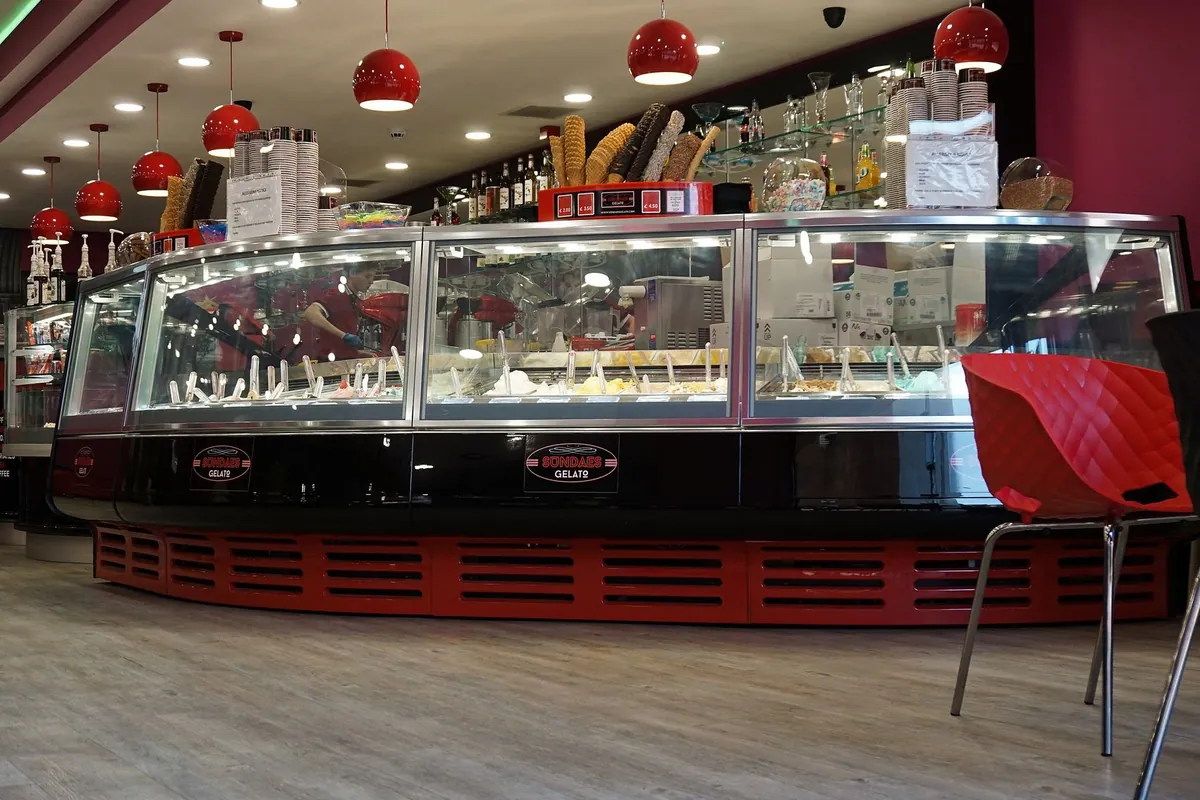 Vetrina curva di una gelateria con le vaschette in vista, i coni impilati sopra e le lampade rosse appese al soffitto