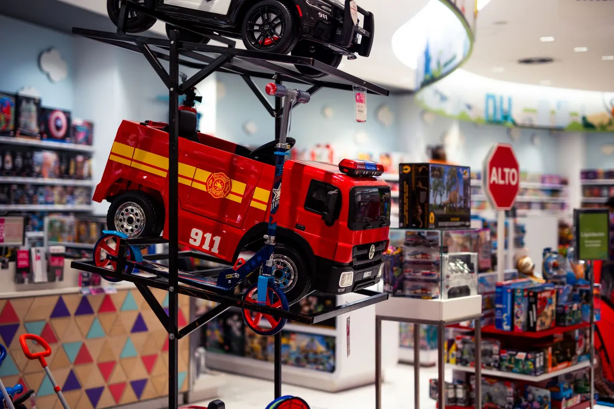 Toy shop with a ride-on fire engine and police car on a metal display rack, scooters underneath and shelves of boxed toys behind