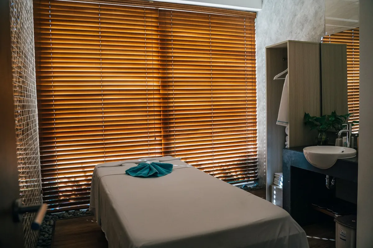 Cabine de massagem com a maca arrumada e uma toalha dobrada em cima, a persiana de madeira cobrindo a parede e a pia ao lado