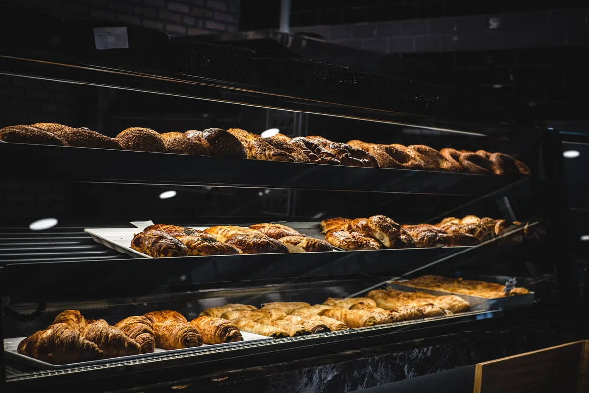 Vitrina de panadería iluminada con tres bandejas: croissants y napolitanas abajo, hojaldres rellenos en el centro y hogazas trenzadas con sésamo arriba