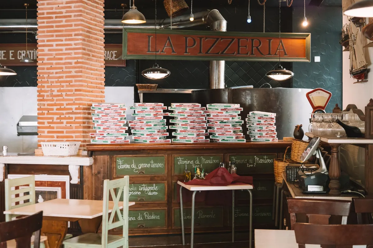 Interior de pizzería con el horno de leña al fondo, torres de cajas para llevar sobre el mostrador y los cajones de harina rotulados a tiza