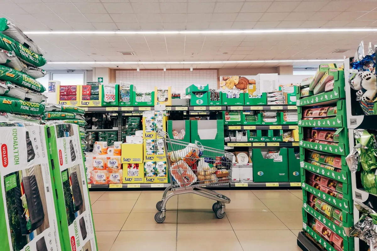 Allée de supermarché avec un caddie plein au milieu, des têtes de gondole chargées et des étiquettes de prix jaunes sur chaque rayon