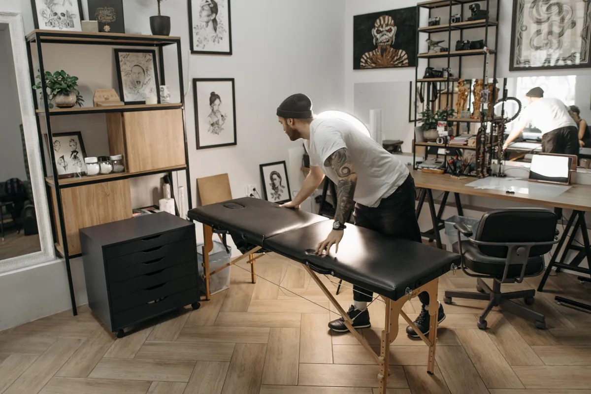 Studio de tatouage avec les murs couverts de dessins encadrés et un tatoueur en bonnet qui prépare la table noire