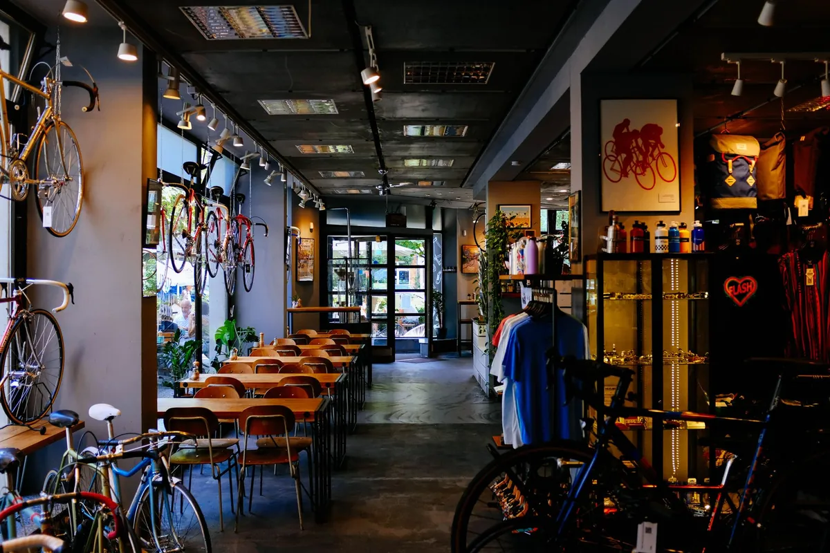 Negozio di biciclette con le bici appese alla parete, tavolini da caffetteria su un lato e l'espositore di maglie e borracce sull'altro