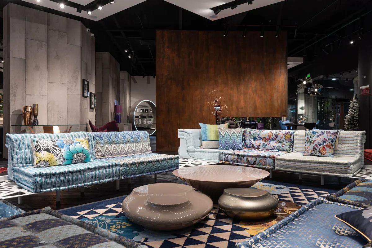 Showroom de uma loja de móveis com dois sofás modulares em tecidos estampados, mesas baixas esculturais e tapetes sobrepostos