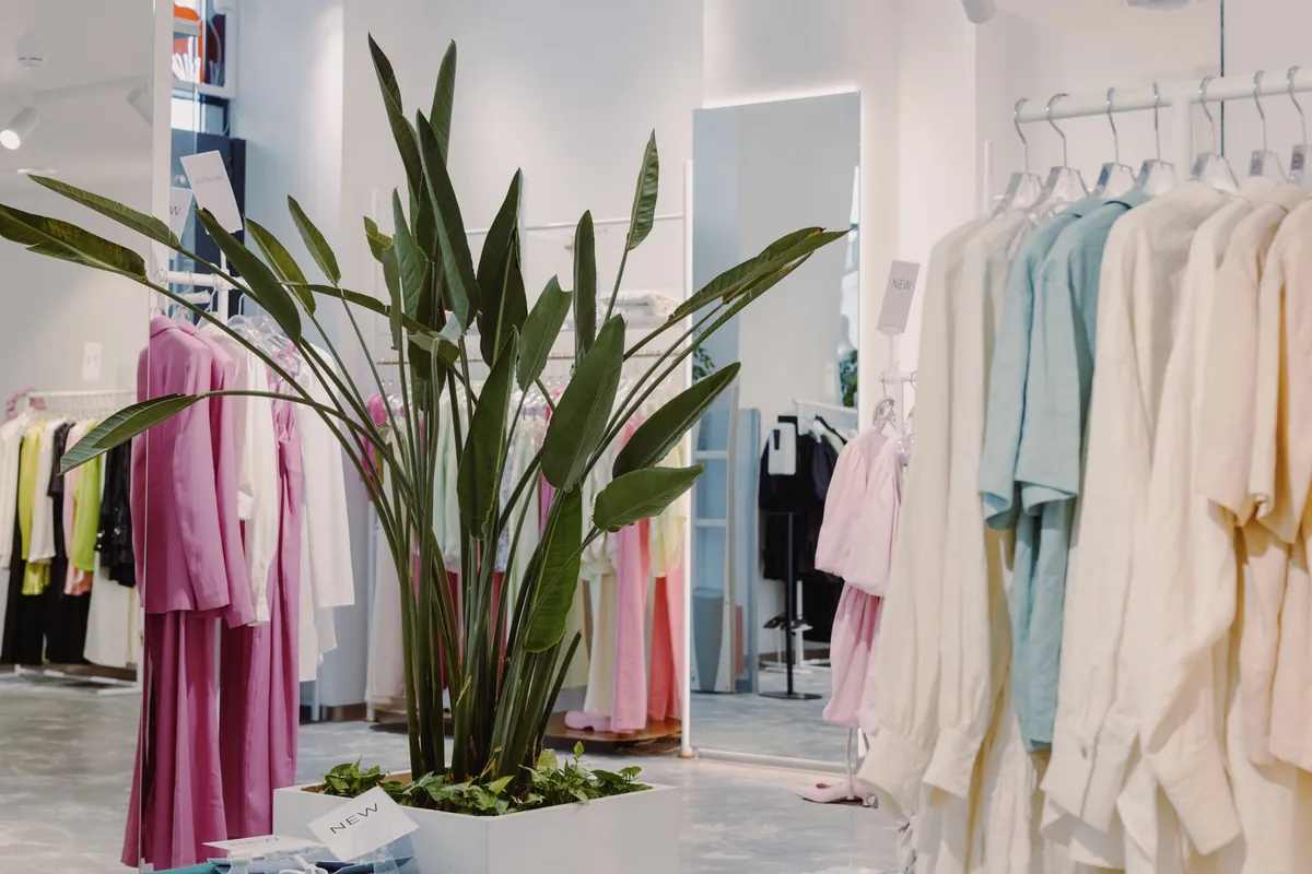 Interior de uma loja de roupas com as araras de peças em tons pastel, um espelho de corpo inteiro ao fundo e uma planta grande no centro
