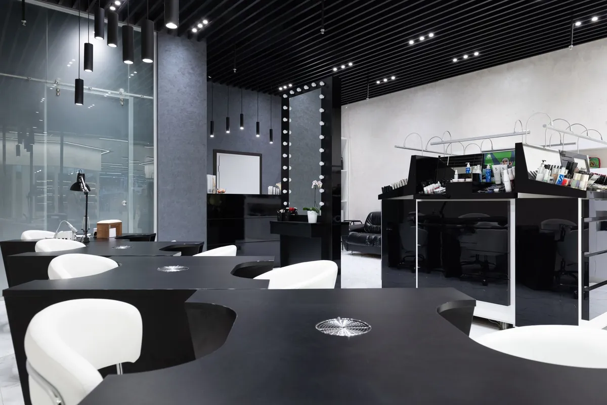 Onglerie avec des tables noires et des chaises blanches, un miroir à ampoules au fond et un écran allumé derrière le présentoir de produits
