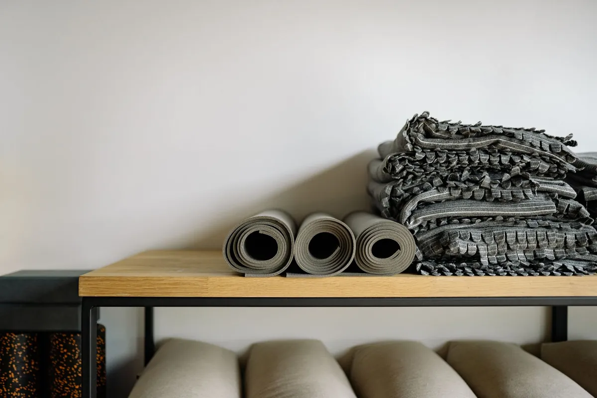 Shelf in a yoga studio with three rolled mats, folded blankets beside them and meditation bolsters on the rack below